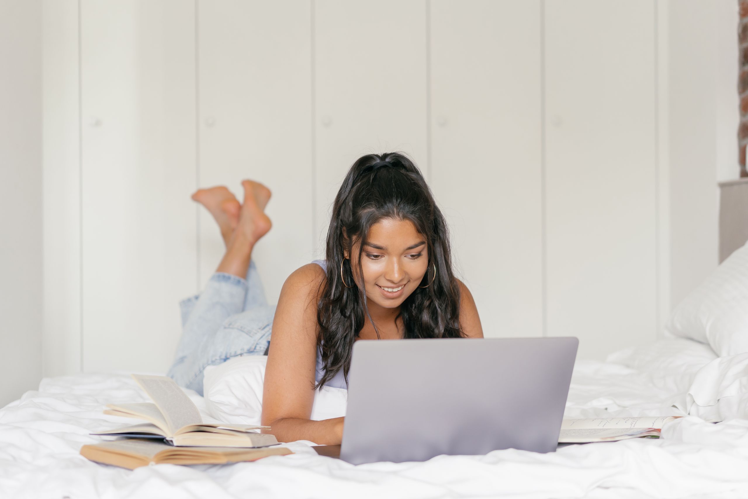 Woman lying on bed with laptop, studying and reading books in a cozy bedroom.